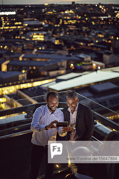 Schrägaufnahme von fröhlichen Geschäftsleuten  die sich mit einem Smartphone selbstständig machen  während sie nach der Arbeit auf der Terrasse stehen und beleuchtet sind