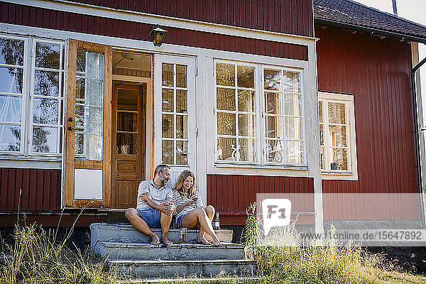Ehepaar betrachtet Smartphone  während es auf Stufen vor dem Blockhaus sitzt