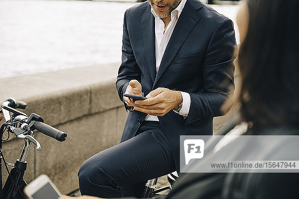 Mittelsektion eines Geschäftsmannes  der ein Mobiltelefon benutzt  während eine Geschäftsfrau auf dem Fahrrad sitzt