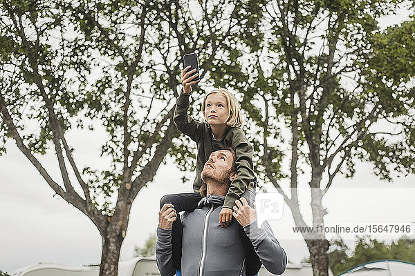 Girl holding mobile phone while sitting on father's shoulders against trees