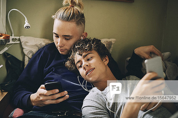 Teenage friends using mobile phone while leaning on bed at home