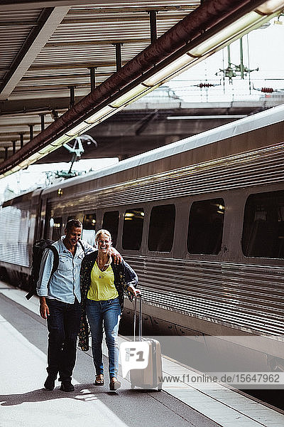 Das glückliche Paar in voller Länge mit Gepäck geht mit dem Zug auf dem Bahnsteig des Bahnhofs