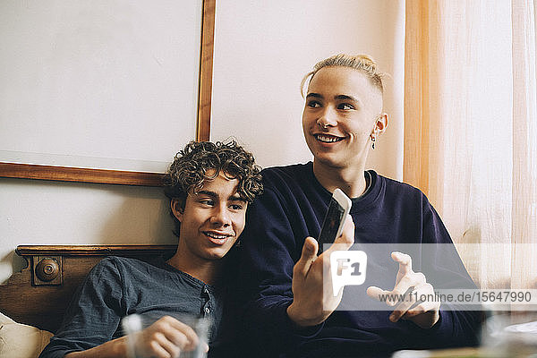 Lächelnder Teenager mit einem Freund  der ein Smartphone zeigt  während er zu Hause wegschaut