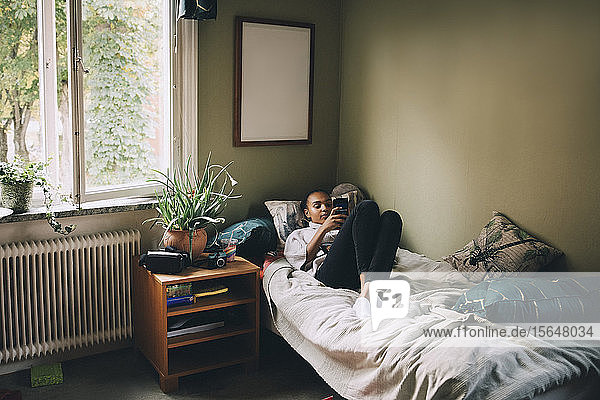 Teenager-Mädchen in voller Länge benutzt Smartphone  während sie zu Hause im Bett liegt