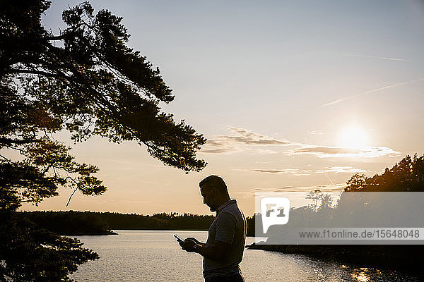 Scherenschnitt eines Mannes mit einem Smartphone  der bei Sonnenuntergang am See steht