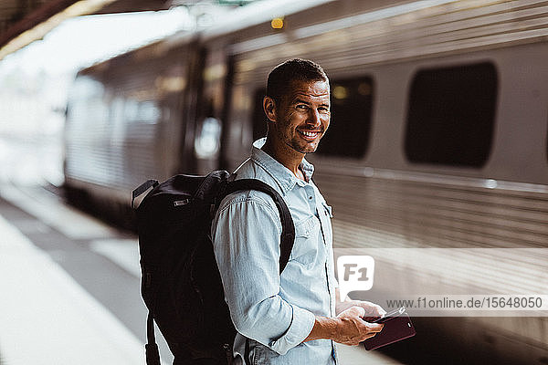 Porträt eines lächelnden Touristen mit Rucksack  der ein Smartphone auf dem Bahnsteig des Bahnhofs hält