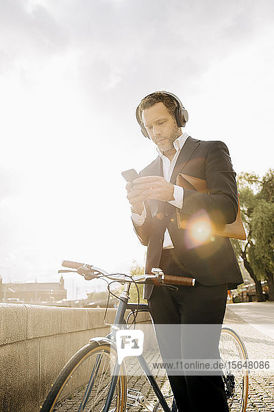 Businessman using mobile phone while standing by bicycle against sky during sunny day