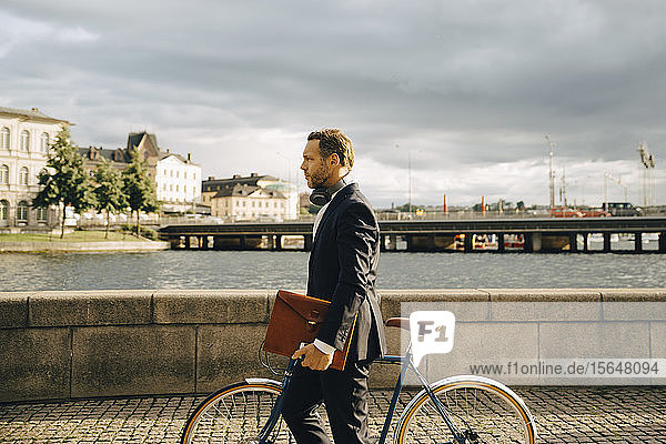 Seitenansicht eines Geschäftsmannes zu Fuß mit dem Fahrrad auf einem Fußweg in der Stadt gegen den Himmel