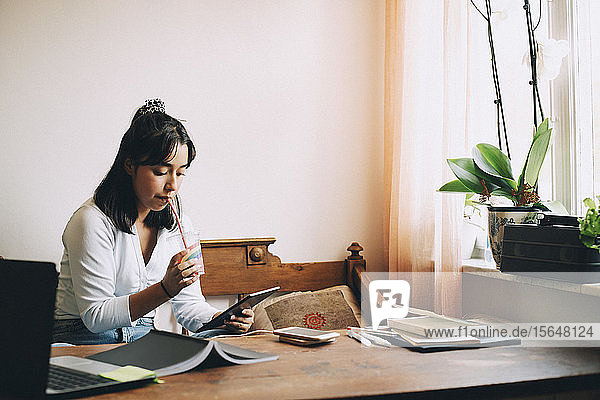 Mädchen trinkt Smoothie  während sie ein digitales Tablet auf dem Sofa zu Hause benutzt