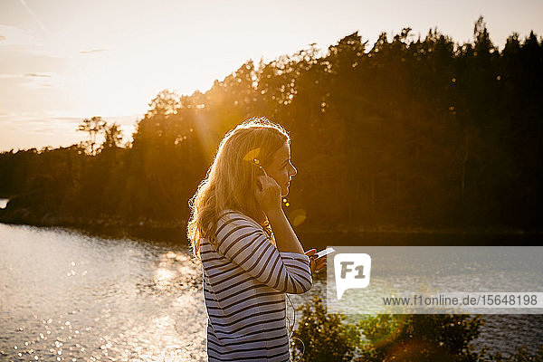 Seitenansicht einer mittleren erwachsenen Frau  die ein Mobiltelefon benutzt  während sie bei Sonnenuntergang am See steht