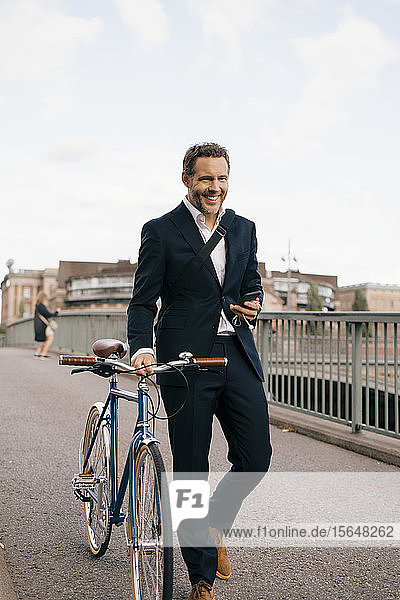 Lächelnder Geschäftsmann hält Smartphone in der Hand  während er mit dem Fahrrad auf der Brücke in der Stadt läuft