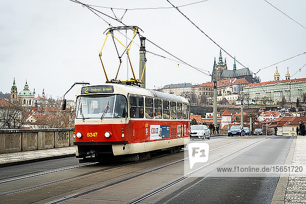 Eine traditionelle rote Straßenbahn fährt über die Manesuv most (Brücke) mit dem Veitsdom und der Prager Burg im Hintergrund  Prag  Tschechische Republik