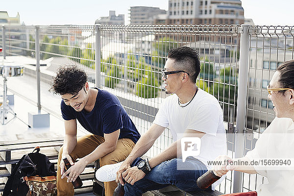 Drei junge japanische Männer sitzen auf einem Dach in einer städtischen Umgebung und trinken Bier.