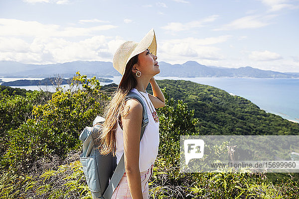 Japanische Frau mit Hut und Rucksack auf einer Klippe stehend im Hintergrund das Meer.
