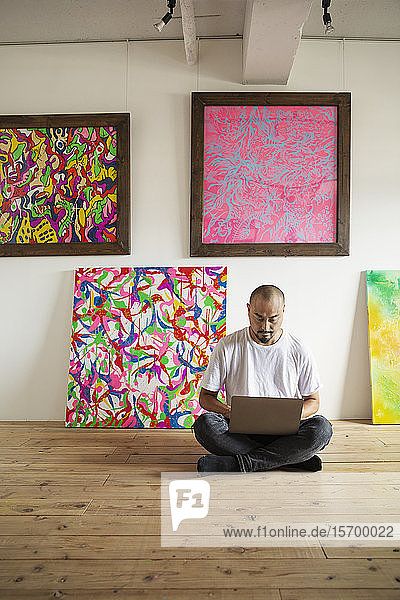 Japanischer Mann sitzt auf dem Boden einer Kunstgalerie und arbeitet an einem Laptop-Computer.