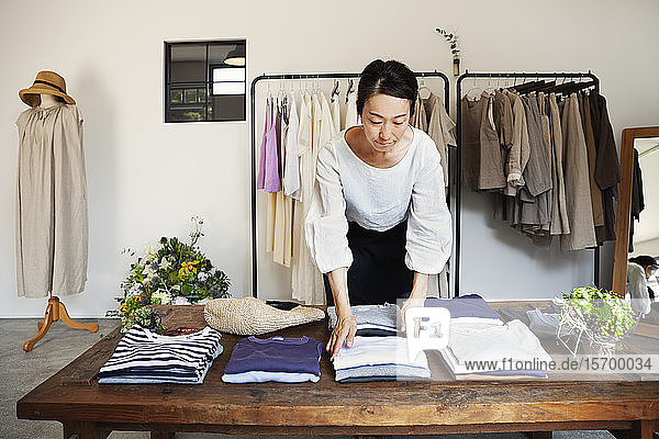 Japanerin  die in einer kleinen Modeboutique steht und T-Shirts auf einem Couchtisch arrangiert.