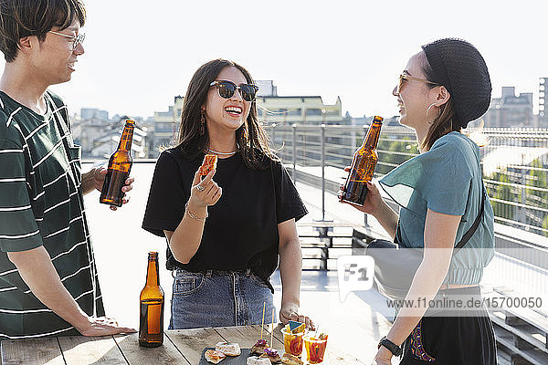 Young Japanese man and two women standing on a rooftop in an urban setting  drinking beer.