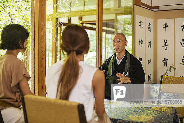 Zwei Japanerinnen und ein buddhistischer Priester knien im buddhistischen Tempel und unterhalten sich.