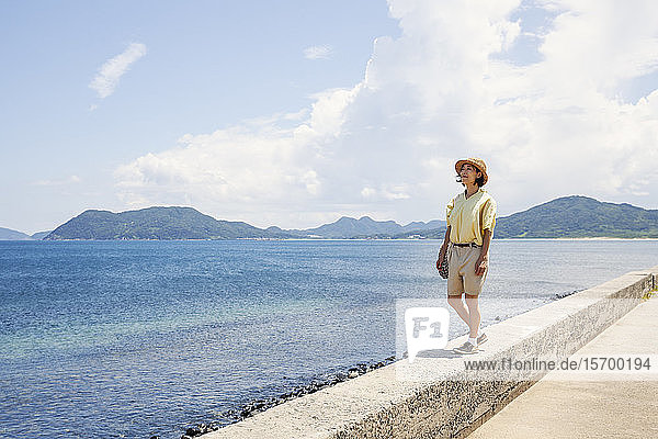 Japanische Frau mit Hut  die an einer Wand steht und auf den Ozean schaut.
