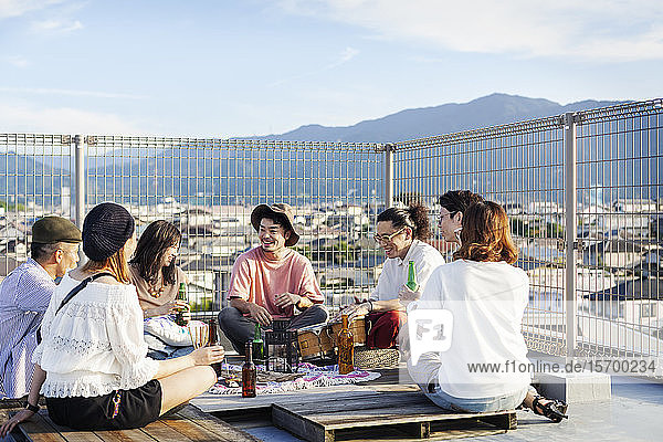 Gruppe junger japanischer Männer und Frauen  die auf einem Dach in einer städtischen Umgebung sitzen und Bier trinken.