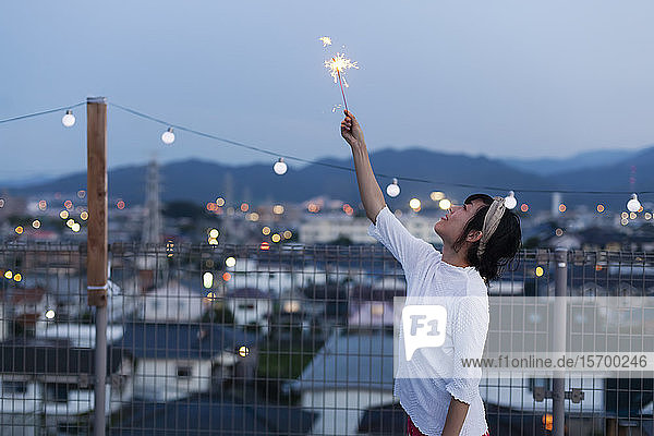Lächelnde junge Japanerin mit einer Wunderkerze in der Hand auf einem Dach in einer städtischen Umgebung.