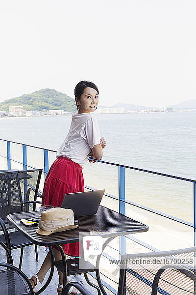 Japanische Fachfrau steht auf dem Balkon eines Arbeitsraumes  Laptop-Computer auf dem Tisch.