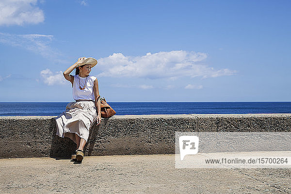 Japanische Frau mit Hut  die an einer Wand sitzt  im Hintergrund das Meer.