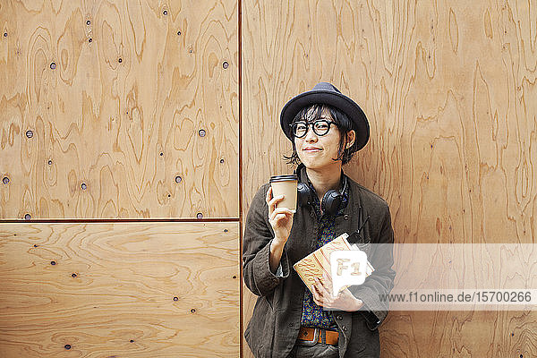Japanese woman wearing glasses and hat standing outside Eco Cafe  holding paper cup and notebook.