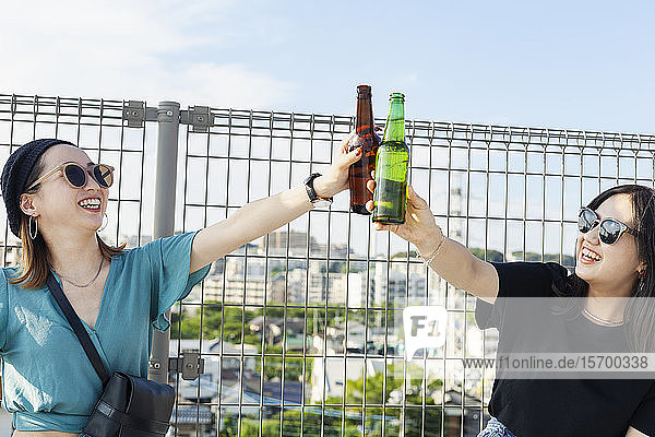 Zwei junge Japanerinnen sitzen auf einem Dach in einer städtischen Umgebung und trinken Bier.