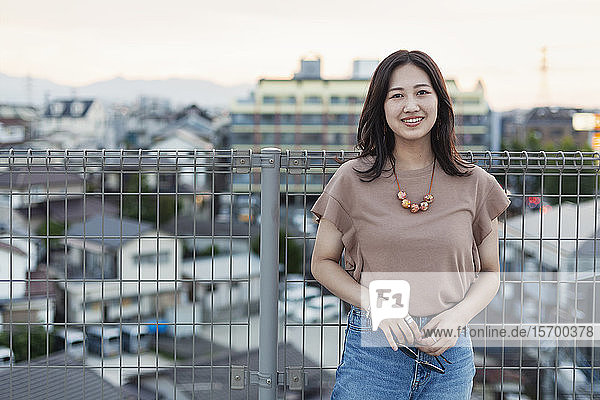 Junge Japanerin  die auf einem Dach in einer städtischen Umgebung steht und in die Kamera schaut.