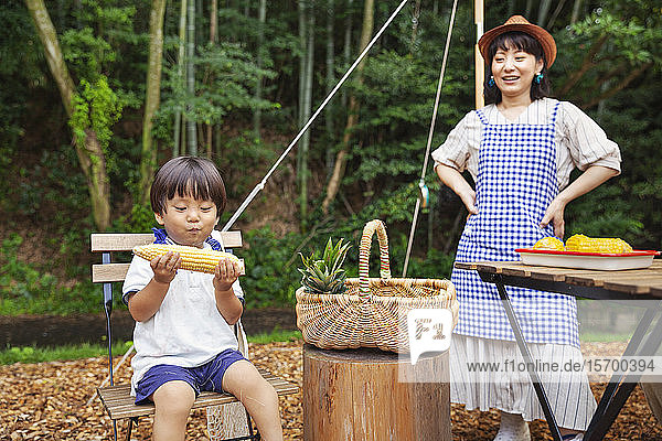 Japanerin steht im Freien  trägt Hut und Schürze  der Junge sitzt auf einem Stuhl und isst Maiskolben.