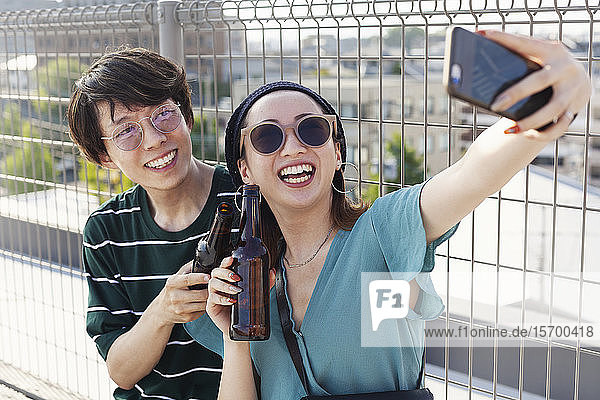 Young Japanese man and woman sitting on a rooftop in an urban setting  taking selfie with mobile phone.