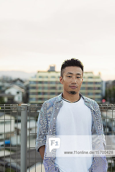 Ein junger Japaner steht auf einem Dach in einer städtischen Umgebung und schaut in die Kamera.