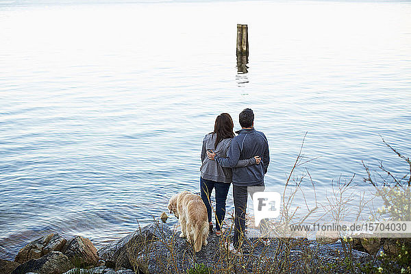 Zärtliches Paar mit Hund  das sich umarmt und den Seeblick genießt