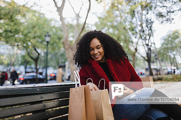 Smiling young woman looking in shopping bags on park bench