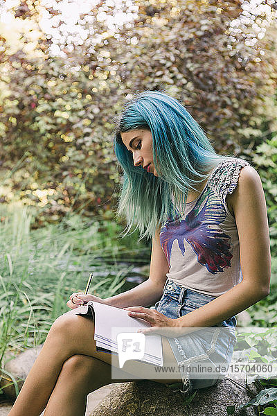 Junge Frau mit blauem Haar schreibt im Park in ein Tagebuch