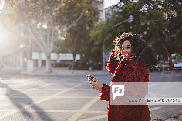 Porträt einer selbstbewussten  lächelnden jungen Frau mit Smartphone auf einer sonnigen Straße in der Stadt