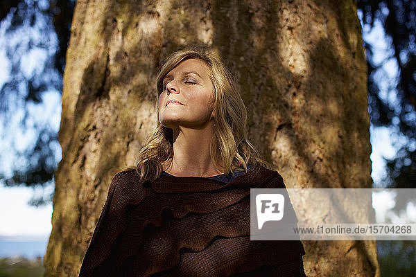 Porträt heitere Frau mit geschlossenen Augen an sonnigem Baumstamm stehend