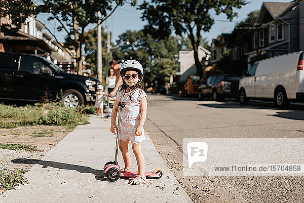 Toddler riding on push scooter in neighbourhood  family in background