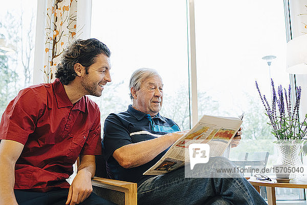 Smiling caregiver sitting by elderly man reading newspaper against window at retirement home
