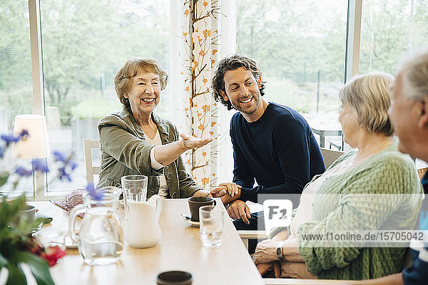 Lächelnde ältere Frau gestikuliert gegenüber Freunden  während sie mit ihrem Enkel im Pflegeheim sitzt