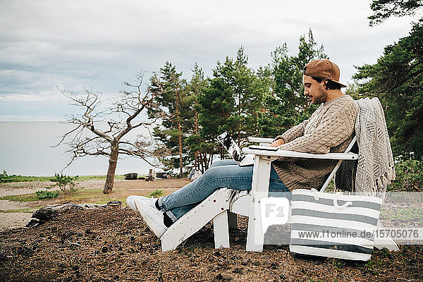 Seitenansicht eines Mannes  der einen Laptop benutzt  während er auf einem Adirondack-Stuhl im Wald sitzt