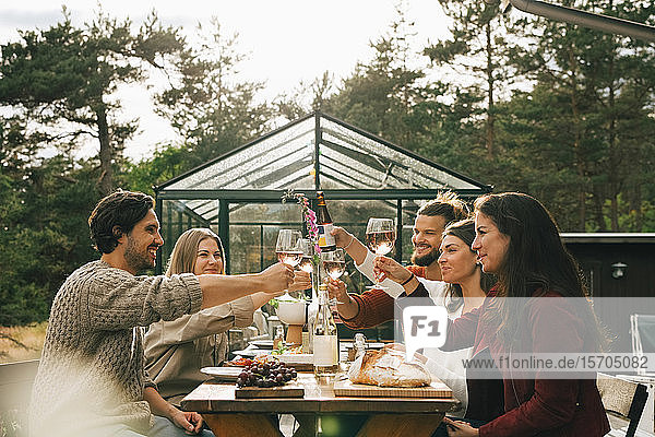 Männliche und weibliche Freunde stoßen auf Weingläser an  während sie auf einer Gartenparty zu Abend essen