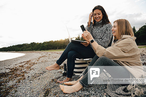 Happy friends looking at smart phone while sitting on shore at beach against sky