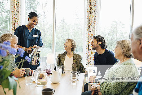 Smiling female nurse serving drink to senior woman sitting with friends and grandson at dining table in nursing home