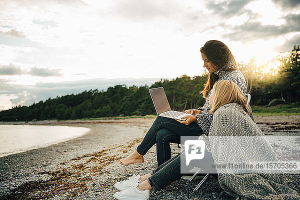 Woman wrapped in blanket looking at friend using laptop on sea shore