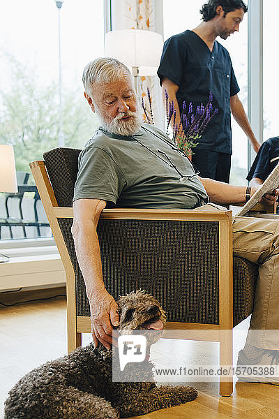 Senioren im Ruhestand sitzen mit Zeitung auf Sessel  während Freunde und Pfleger im Altenpflegeheim ihren Hund streicheln