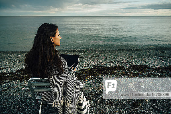 Nachdenkliche Frau mit Laptop sitzt bei Sonnenuntergang auf einem Stuhl am Strand