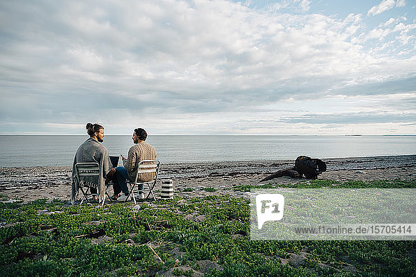 Männliche Freunde diskutieren am Laptop  während sie auf Stühlen am Strand gegen den Himmel sitzen