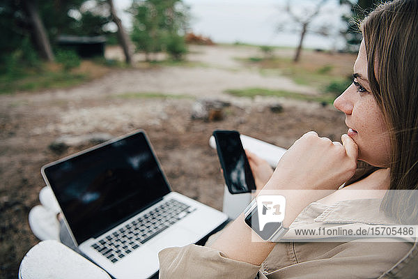 Nachdenkliche Frau mit Smartphone und Laptop sitzt im Freien